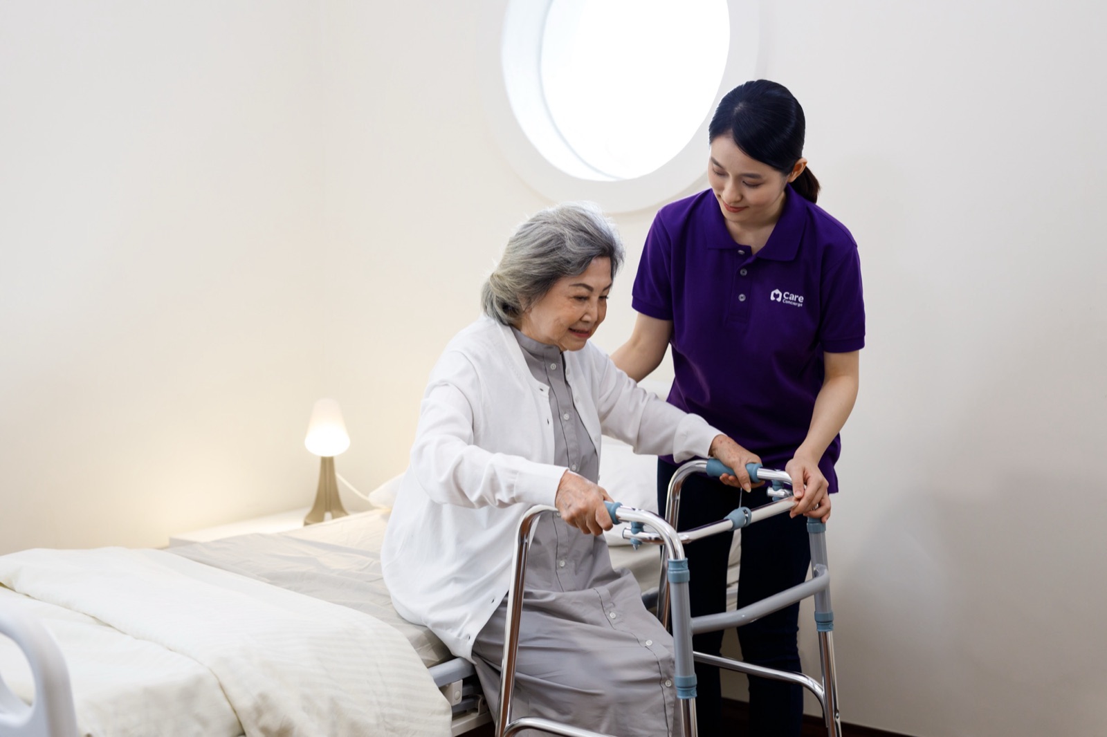 A caregiver steadying an older woman as she stands up from her bed with a walking frame