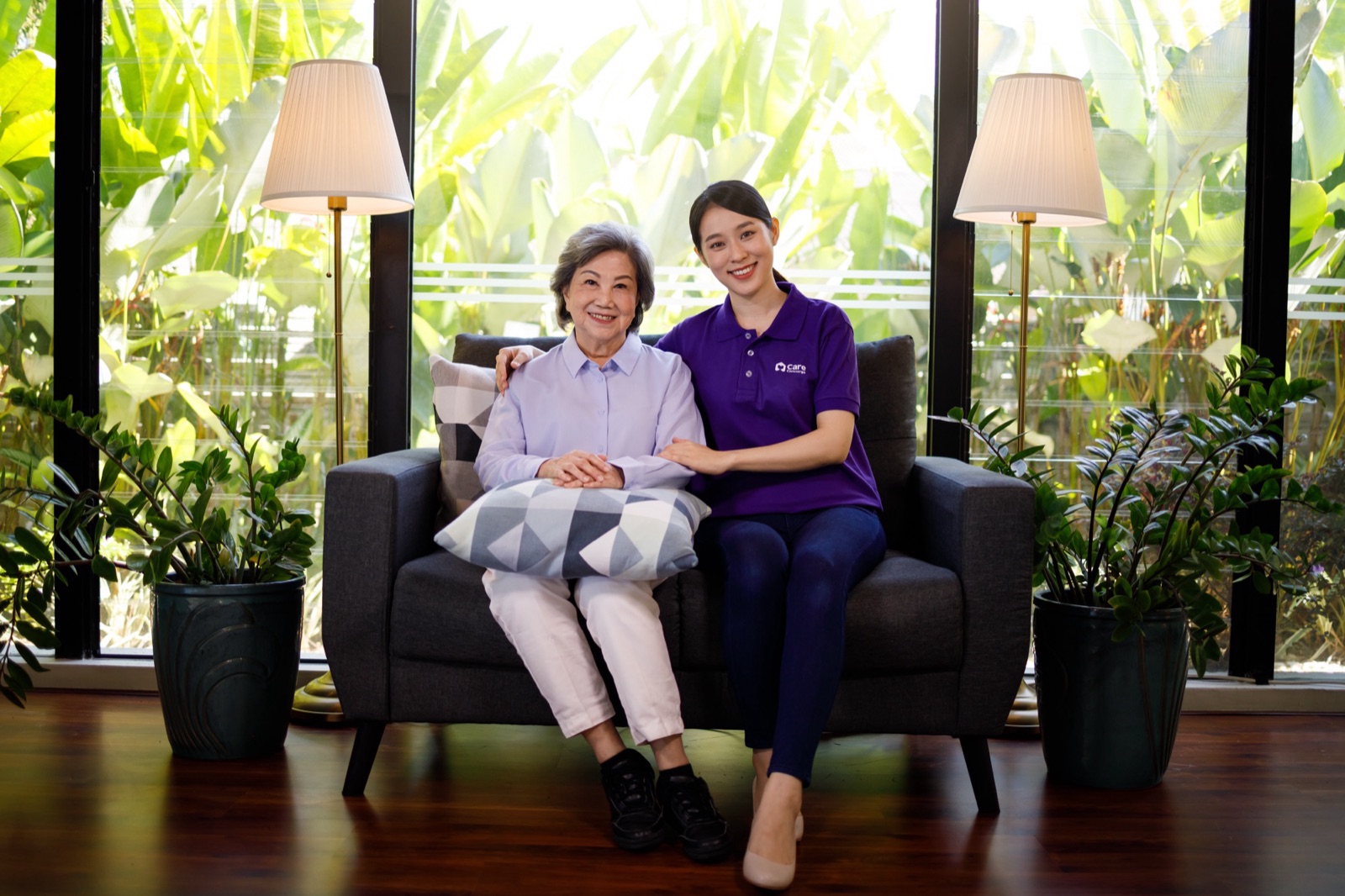 A smiling caregiver in a purple polo sitting beside an older woman on a sofa at home