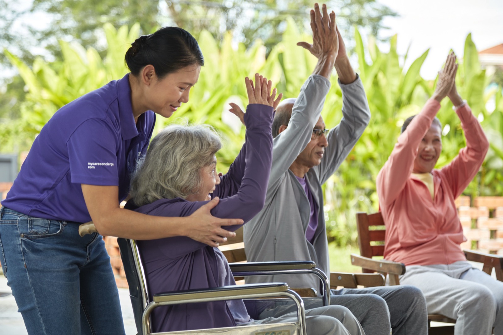 A caregiver helping an older woman in a wheelchair stretch her arms overhead, beside two seniors doing the same outdoors